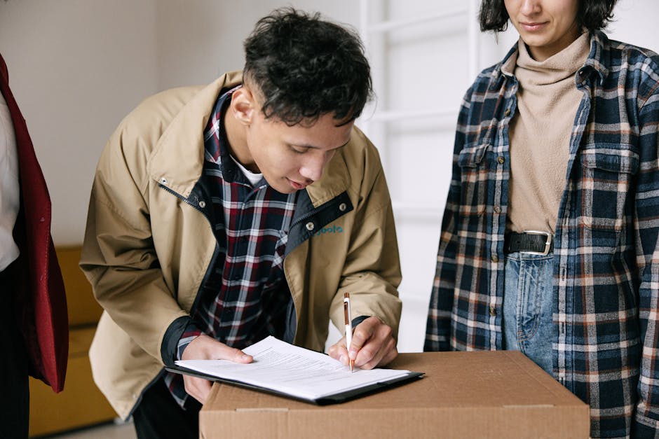 A young man with dark, curly hair dressed in a beige jacket over a checked shirt is seen signing a document placed on top of a cardboard box. Standing next to him is a woman wearing a plaid shirt over a beige turtleneck, with her hands resting on her hips. The scene takes place indoors, possibly in a home or storage area, with a visible door or partition in the background. The cardboard box appears to be part of a home relocation process, as it is likely pre-packed for moving. In the background, there are other boxes and items, indicating an ongoing packing or moving activity. The lighting is bright and natural, emphasizing the details of the individuals and the packing materials. This image supports themes of packing, organisation, and administrative tasks associated with home removals and furniture transport during a move, aligning with services offered by Pimlico Removals.