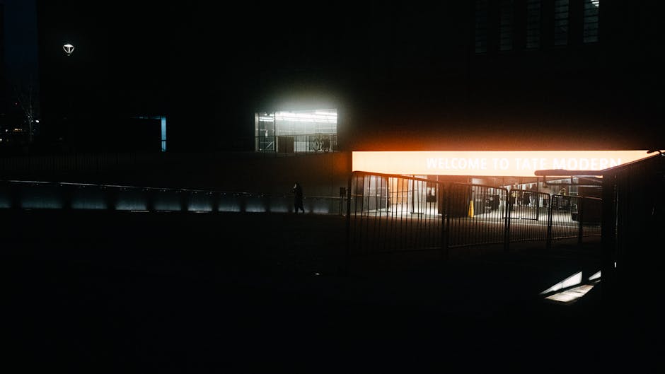 Nighttime exterior view of a modern building with a brightly lit orange sign displaying 'WELCOME TO TATE MODERN' situated above an entrance area. In the foreground, a paved pavement is visible with a metal railing and a few darkly silhouetted figures. To the left, part of a vehicle, possibly a van used for home relocation and furniture transport, is parked adjacent to the building. The building's facade shows a large glass window with interior lighting, and the surrounding area is dark, emphasizing the illuminated signage and entrance. The scene suggests preparation for a house removal or moving service at a prominent cultural venue, with elements indicating logistics such as loading or unloading processes occurring nearby.